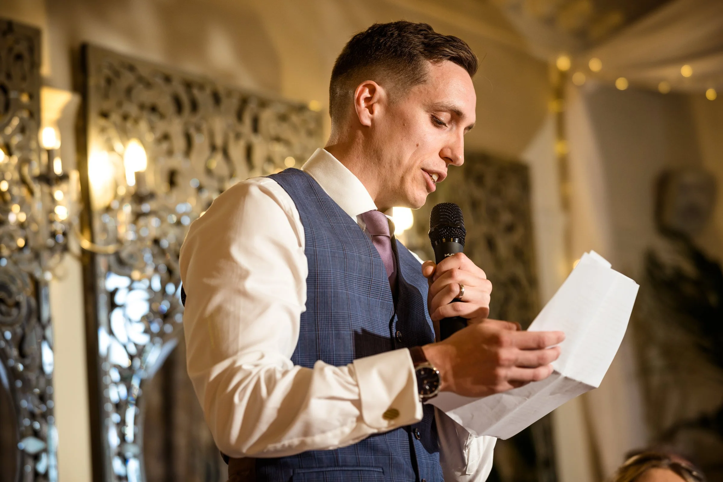 Man in a vest and tie reading from a paper while holding a microphone at an event.
