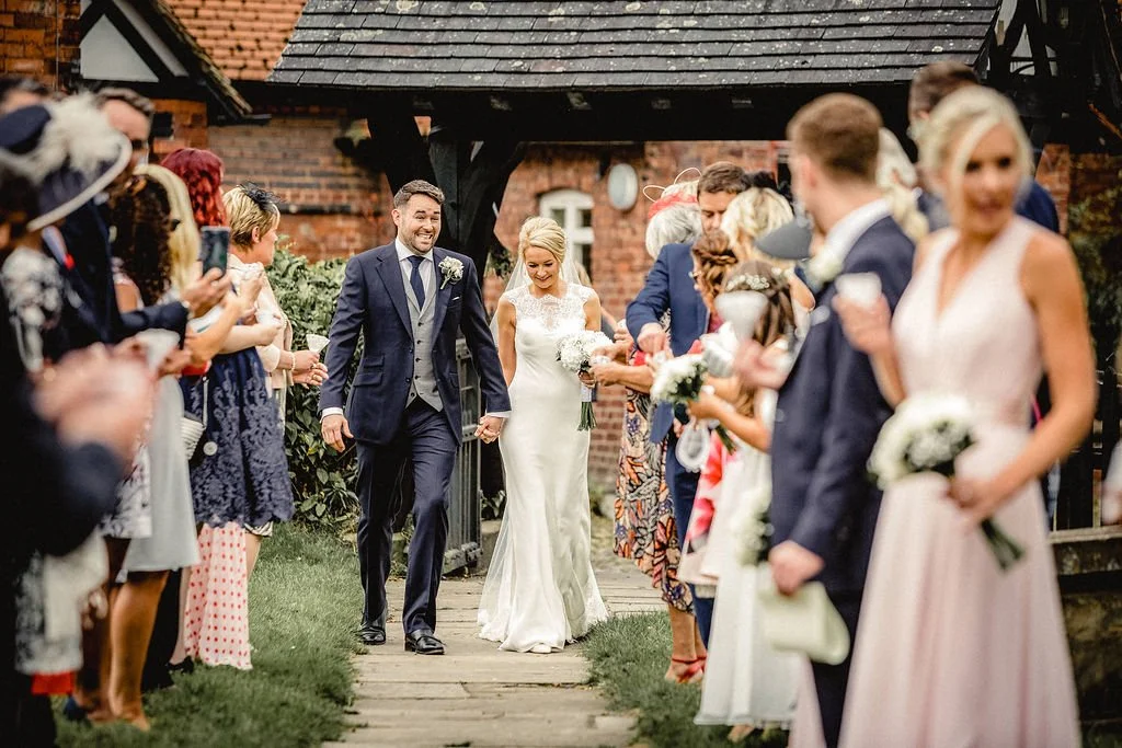 Bride and groom walking hand in hand down aisle at outdoor wedding, surrounded by guests in formal attire, with brick building in background.