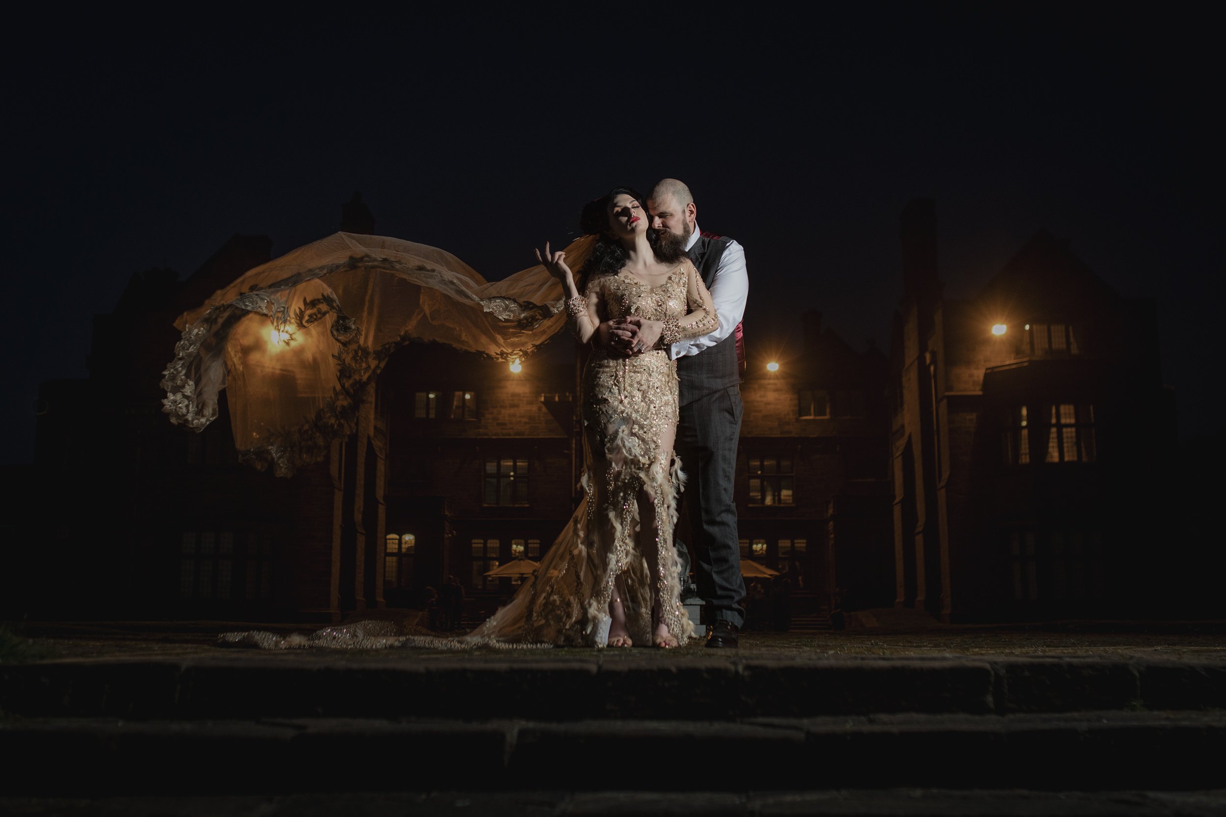 A couple in elegant wedding attire stands close together outdoors at night, with a large gothic-style building and warm lighting in the background.