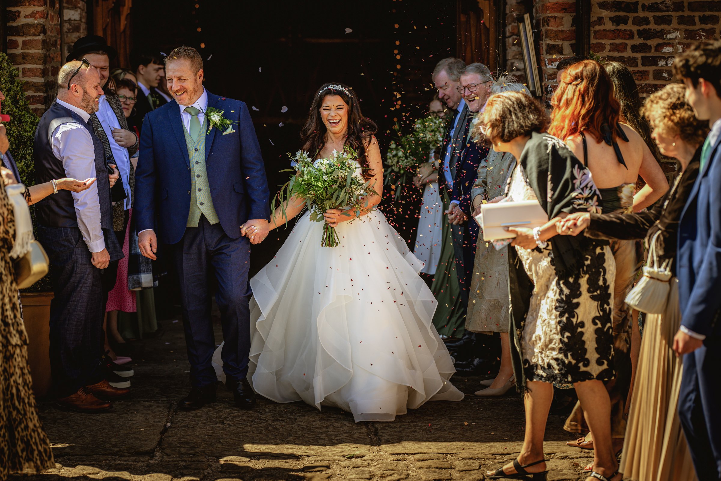 Bride and groom walking hand in hand, smiling, surrounded by guests throwing confetti at a wedding celebration.
