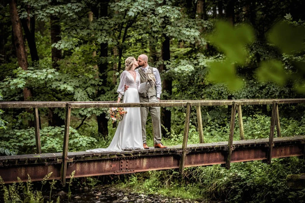 A bride and groom sharing a kiss on a rustic wooden bridge surrounded by lush green trees.