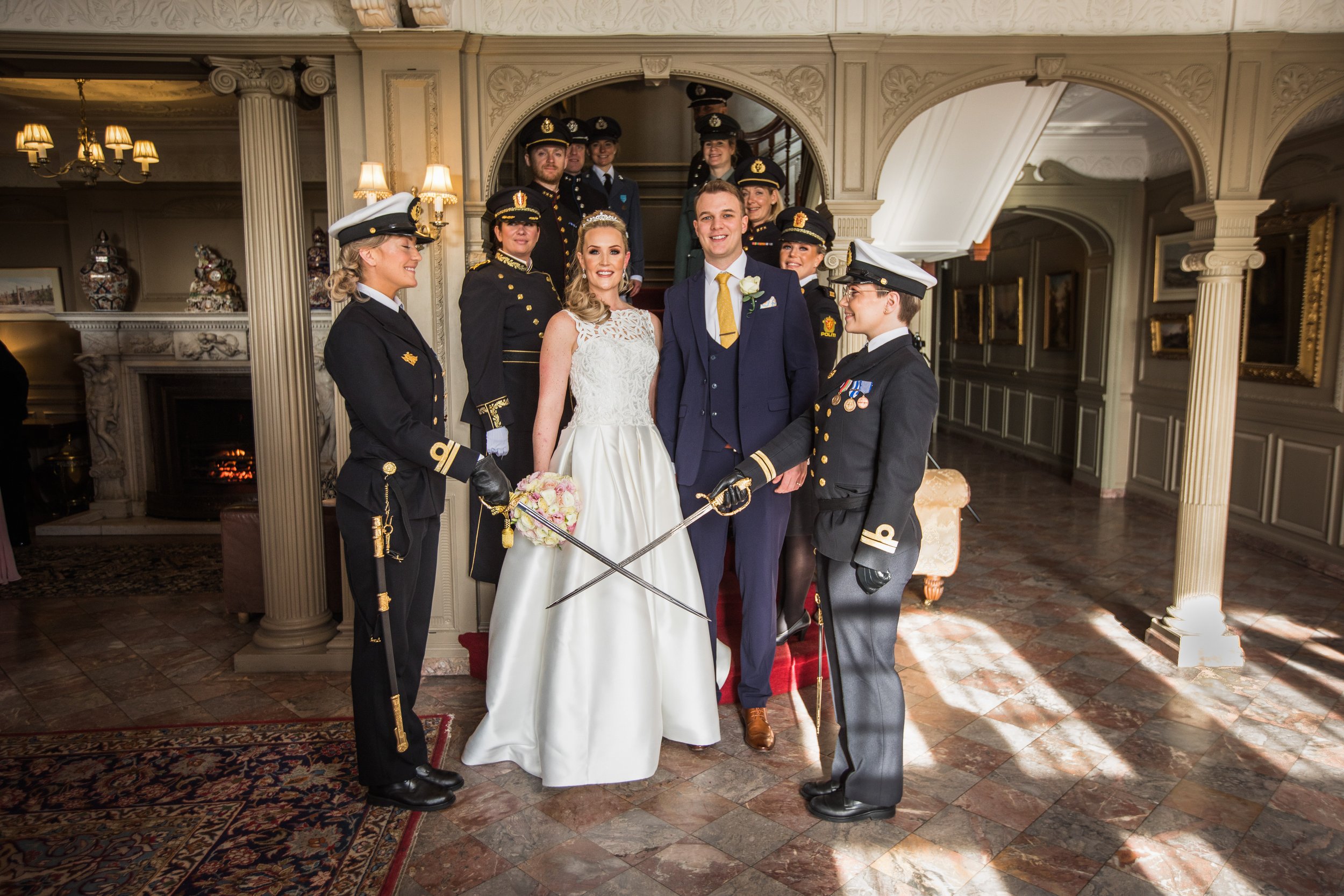 A wedding celebration with the bride and groom in the center, surrounded by military personnel in uniform, inside an elegant, ornately decorated room with a fireplace and vintage furniture.
