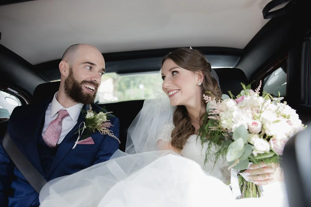 Bride and groom smiling and looking at each other inside a car, with the bride holding a bouquet of flowers.