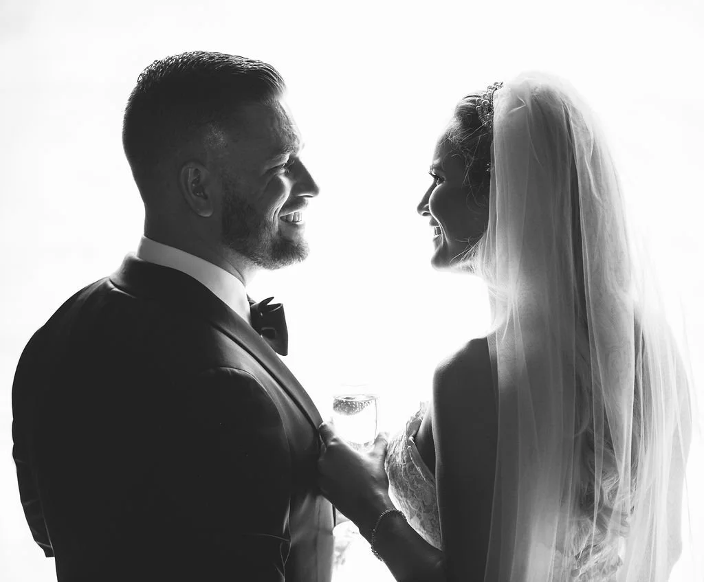 A black and white photo of a bride and groom facing each other, smiling, with the groom holding a drink and wearing a tuxedo, and the bride in a wedding dress with a veil.