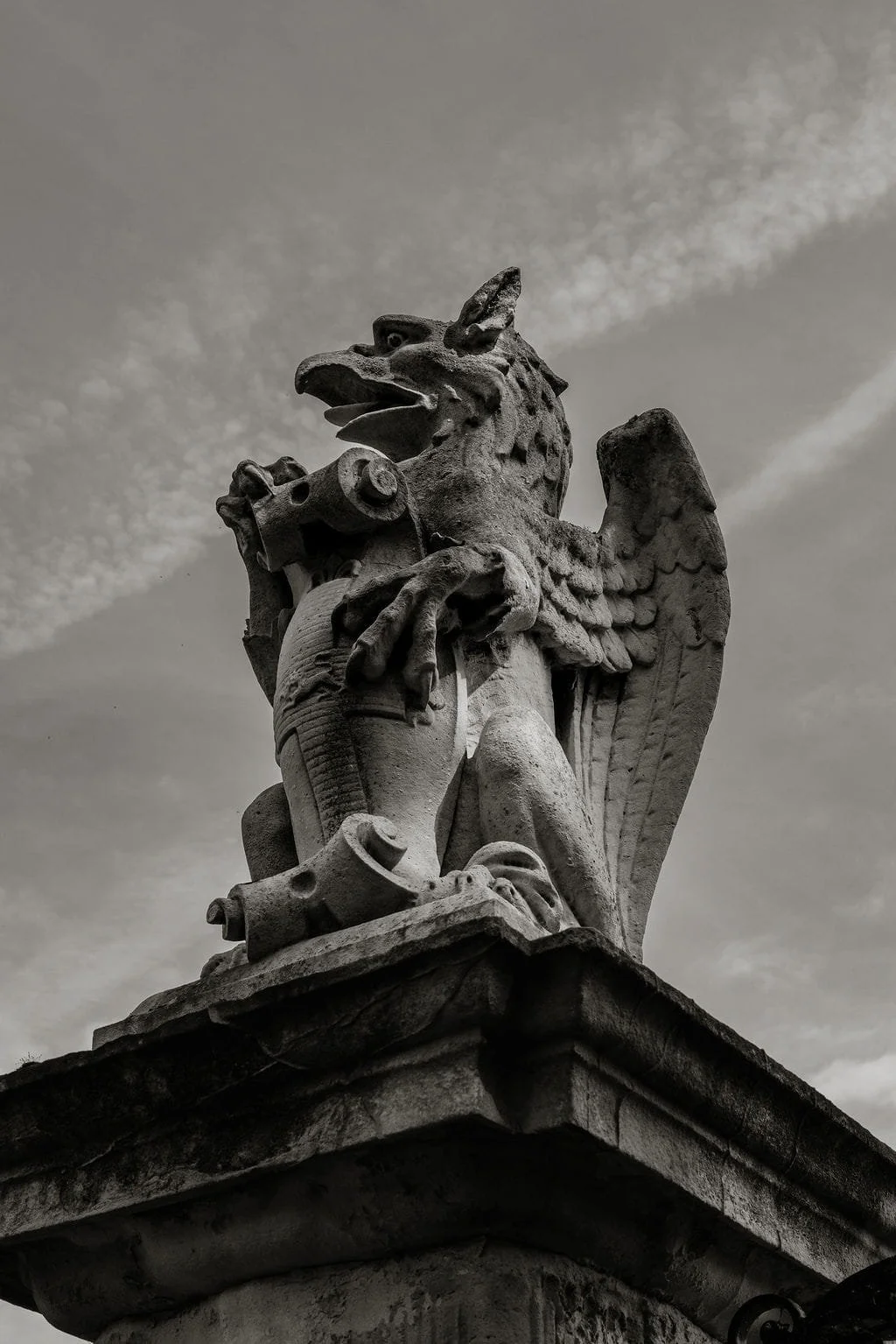 A stone sculpture of a creature with the body of an eagle, head of a wolf, and holding a decorative object, set against a cloudy sky.