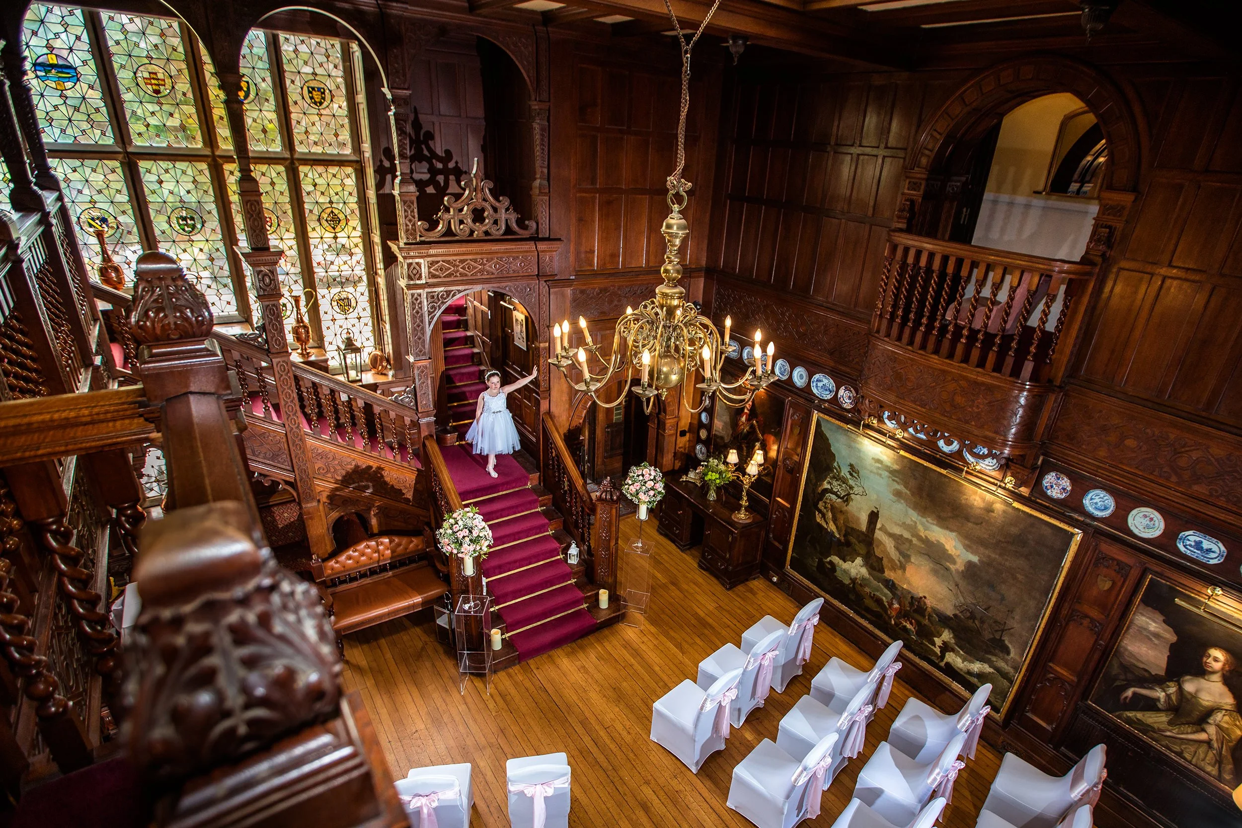 Elegant wooden interior of a historic building with a grand staircase, stained glass windows, chandeliers, and decorated walls. A young girl in a white dress stands on the staircase, and rows of white chairs are set up for a ceremony.