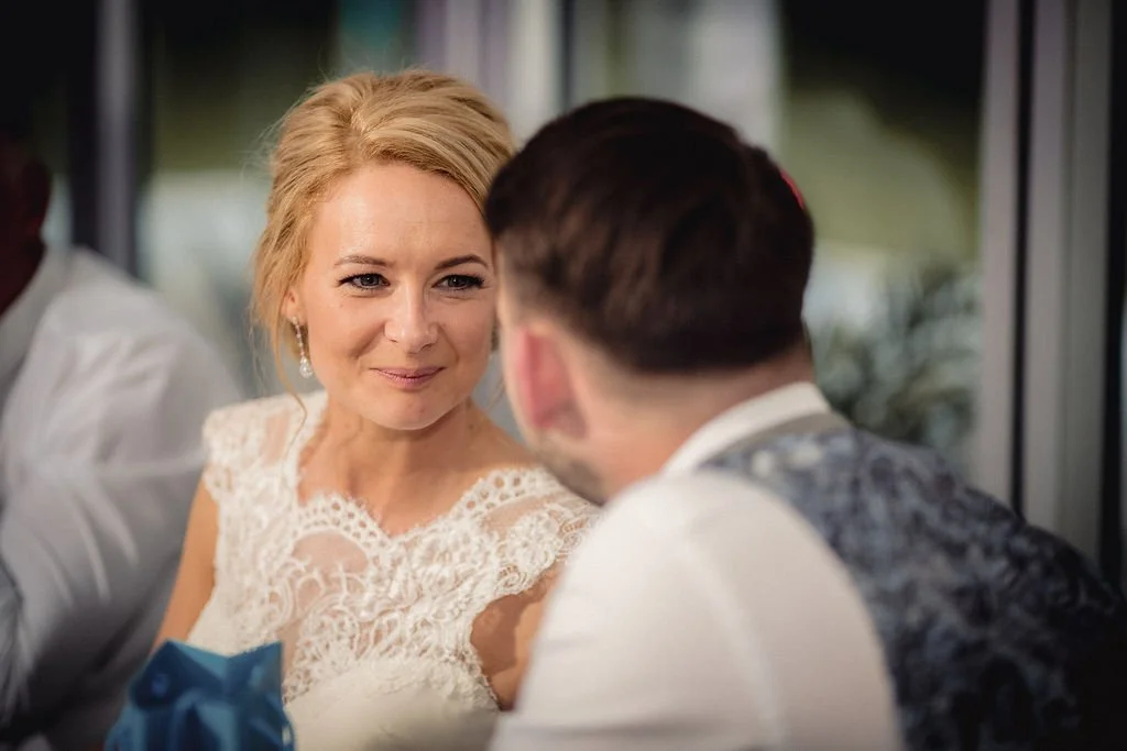 A woman with blonde hair in an elegant lace dress looking at a man with dark hair and a vest, both seated at a table during a formal event.