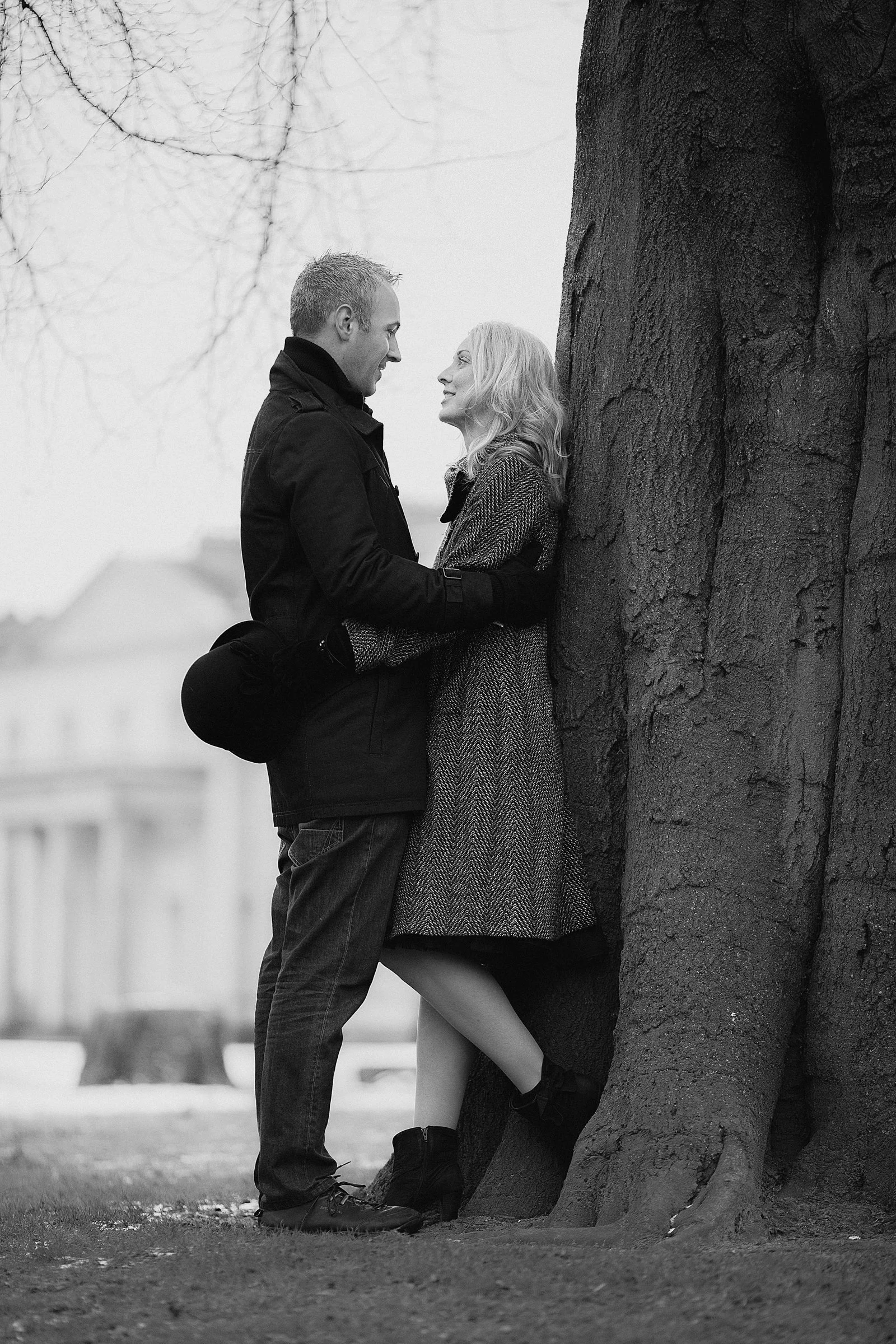 A black and white photo of a man and woman standing close, facing each other, leaning against a large tree. They look at each other with affection. The woman has blonde hair, wears a coat, and the man has short hair, wearing a jacket. The background 