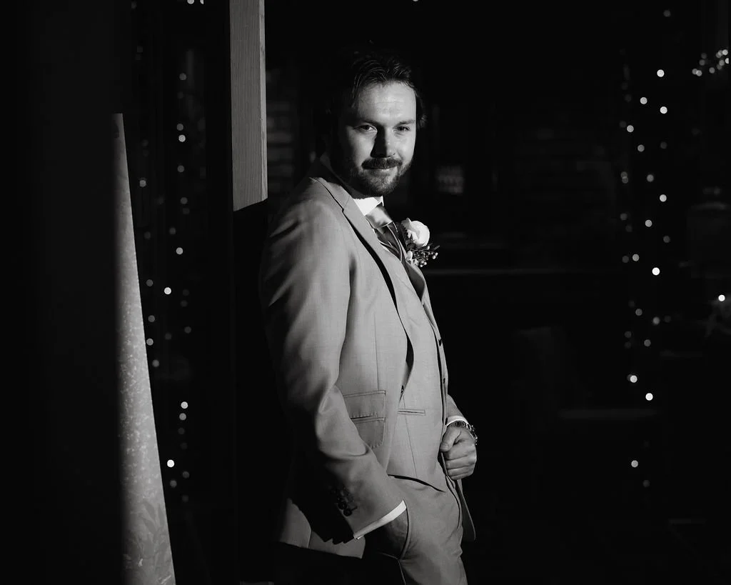 A man in a light-colored suit with a boutonniere, standing indoors at night, looking at the camera with a confident expression.