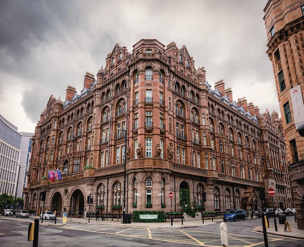 A large, historic red brick building with detailed architecture on a city street, with cars parked and driving by, under a cloudy sky.