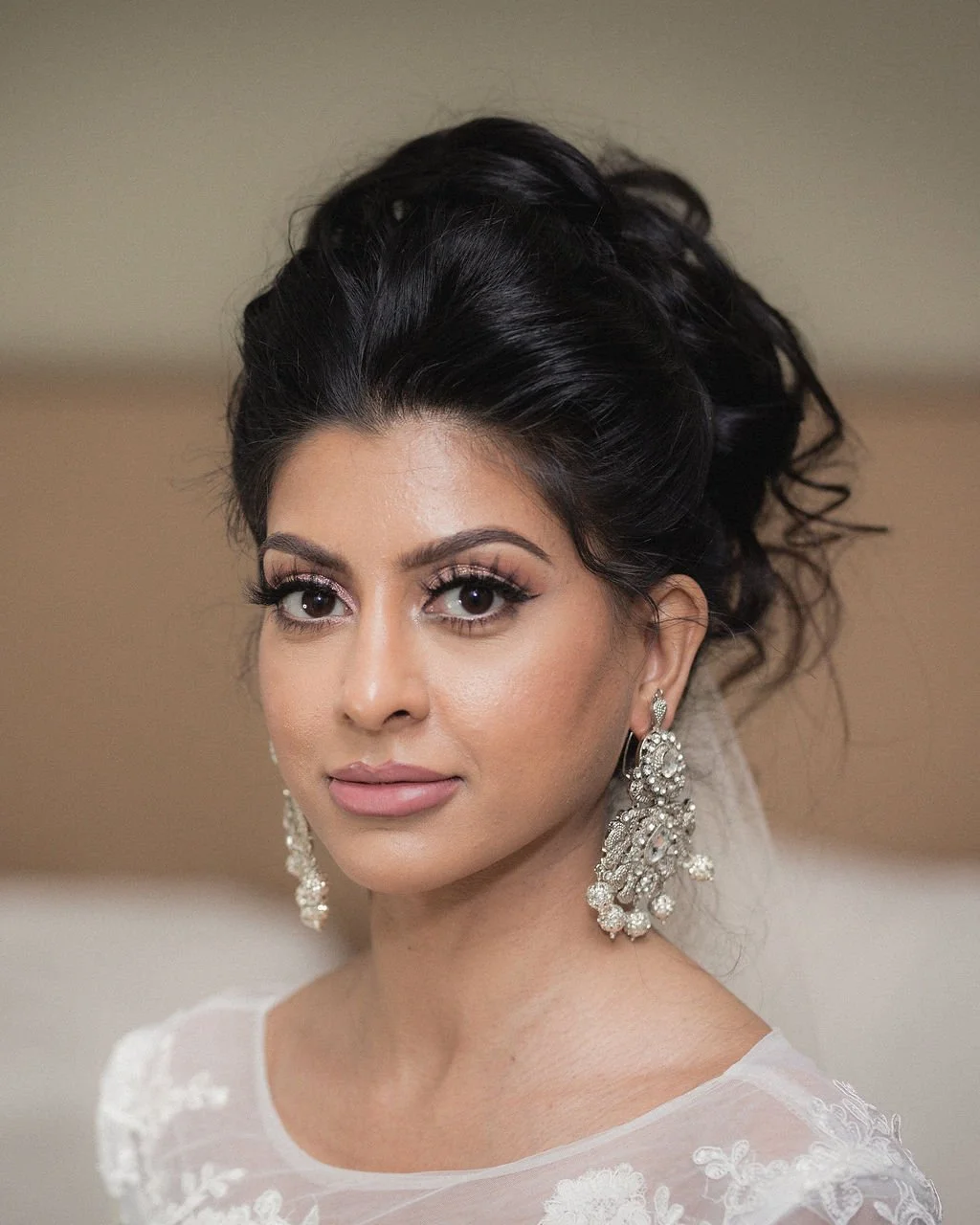 A woman with dark, styled hair and large, ornate earrings, wearing a white lace dress, looking directly at the camera.