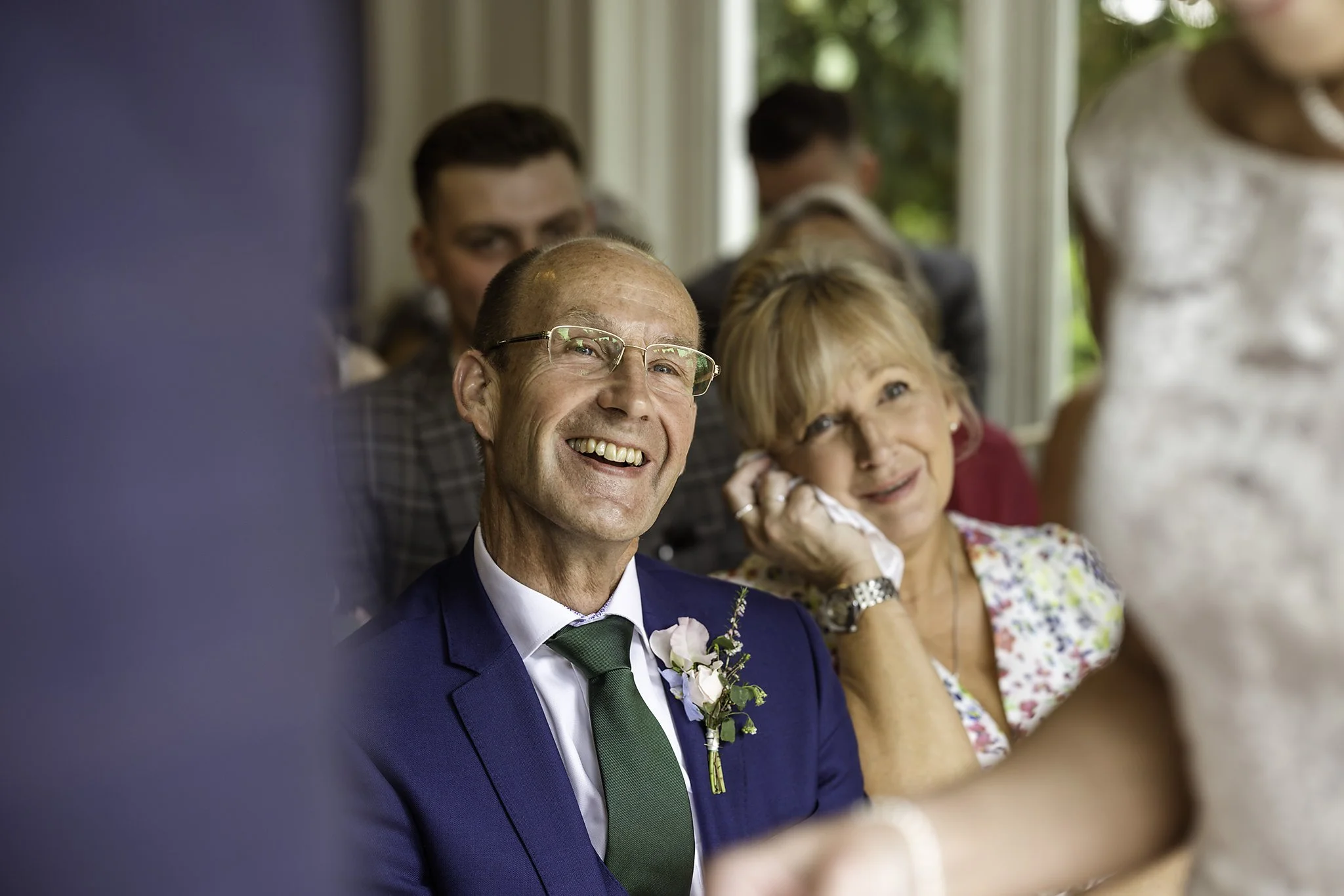Smiling man in a blue suit and glasses sitting next to a woman with short blonde hair, who is touching her face with a tissue, at a wedding or formal event.