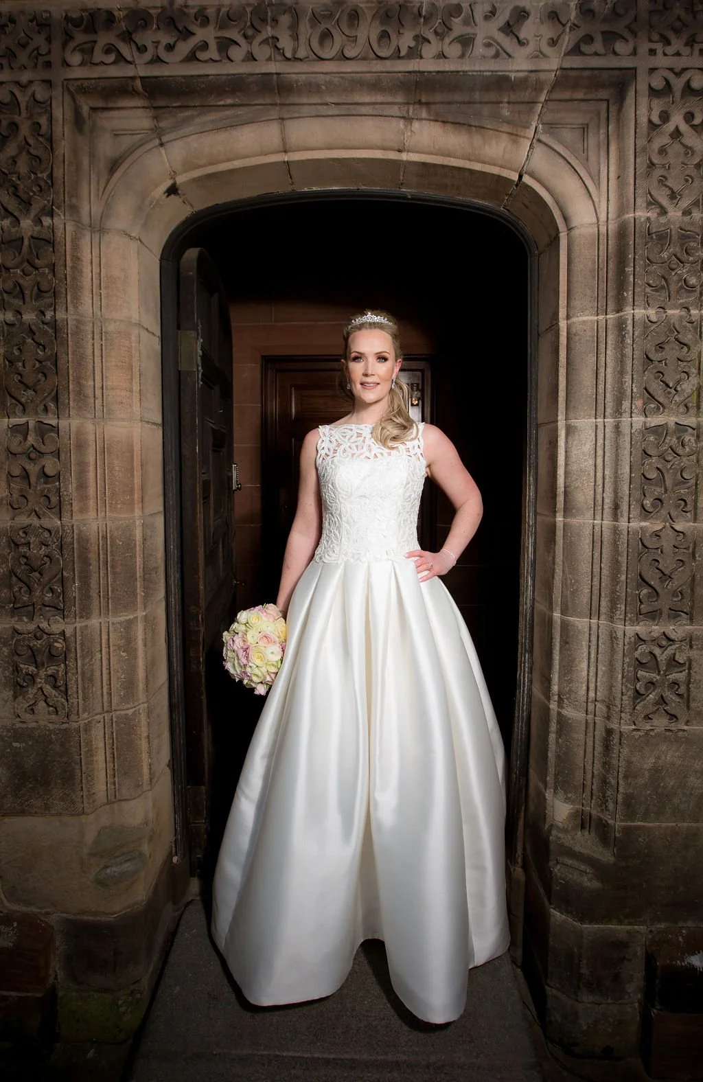 Bride in a white wedding gown holding a bouquet standing in an arched doorway with stone and ornate carvings.