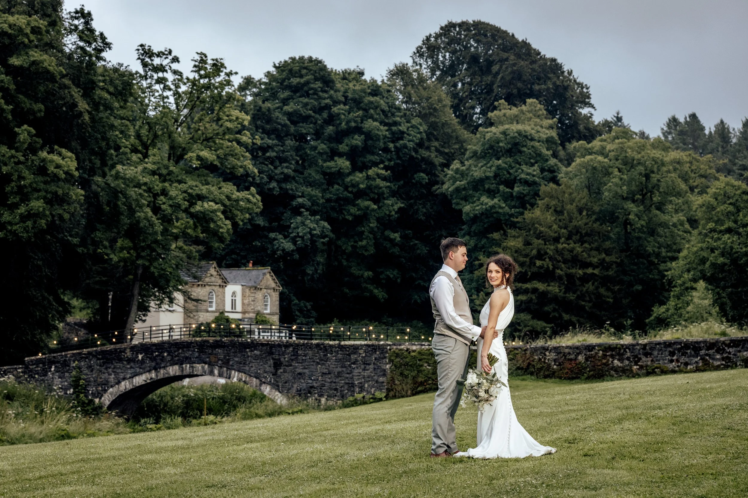 A young couple, possibly newlyweds, standing outdoors on grass, holding hands, with a stone bridge, houses, and lush green trees in the background. The woman is wearing a white dress and holding a bouquet of flowers, while the man is dressed in a ves