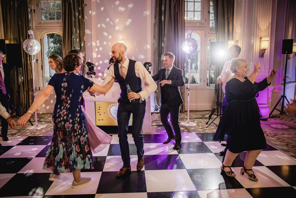 People dancing and enjoying at an indoor wedding reception with a black and white checkered dance floor, disco balls, and purple and white lighting.
