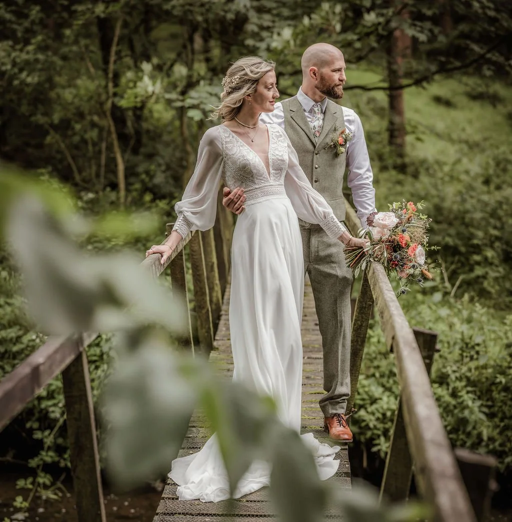 A bride and groom standing on a wooden bridge in a lush green forest, dressed in wedding attire, holding hands, with the bride holding a bouquet of flowers.