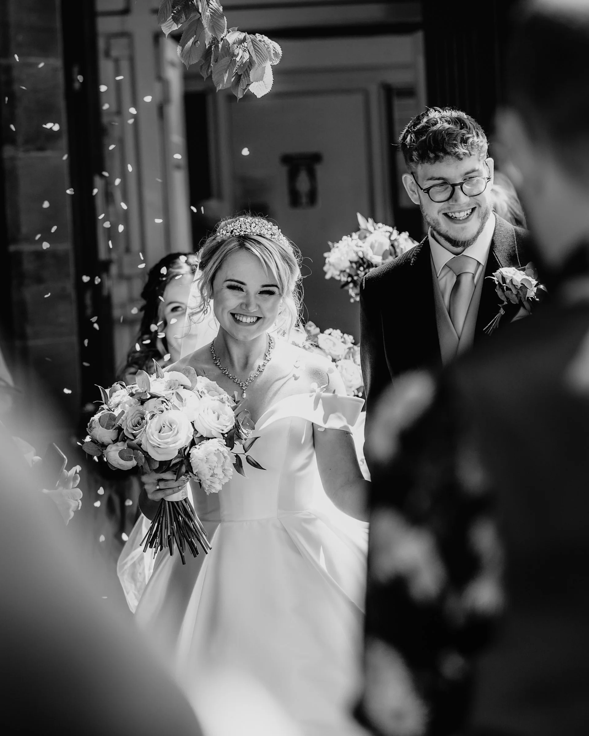 A bride and groom smiling during their wedding celebration, surrounded by guests, with the bride holding a bouquet of flowers.