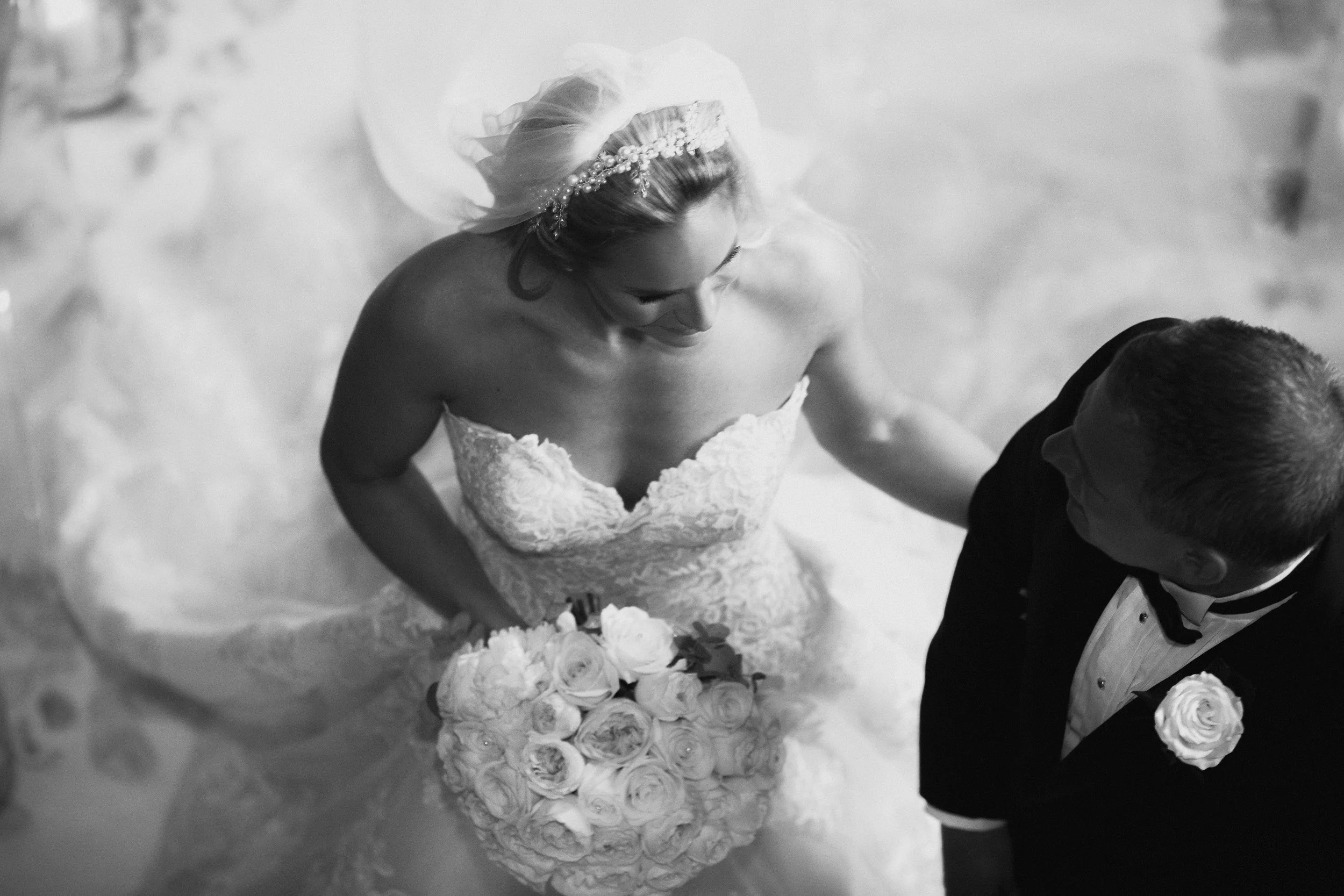 Black and white photo of a bride in a strapless gown holding a bouquet of roses and a groom in a tuxedo with a boutonniere, seen from above.