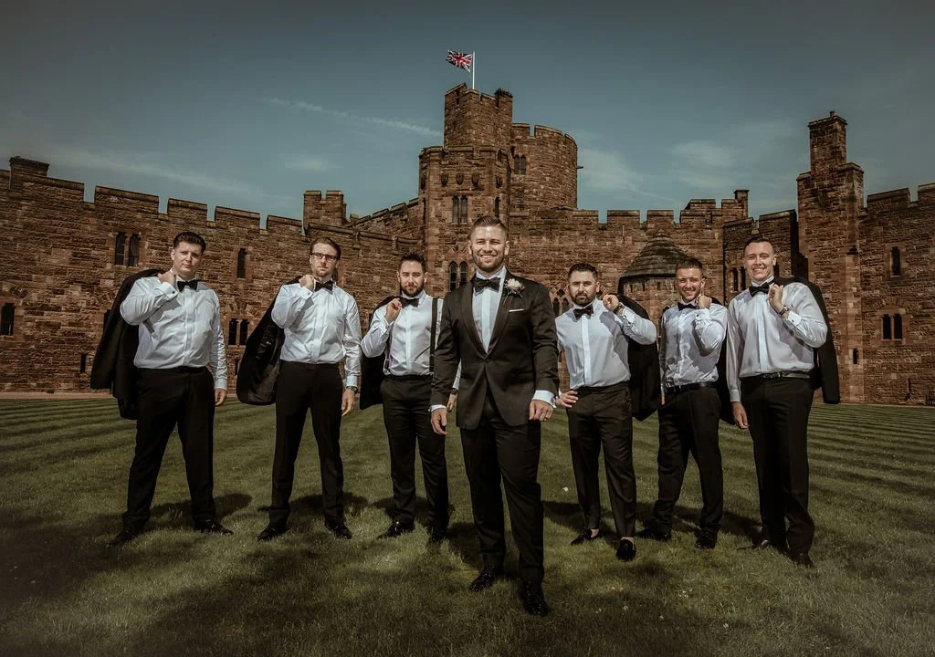 A group of seven men dressed in tuxedos with bow ties, carrying suits jackets over their shoulders, standing in front of a historic castle with a flag on top.