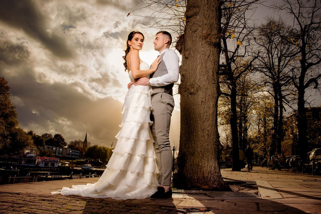 A bride and groom stand close to each other outdoors on a paved walkway next to a large tree, with a cloudy sky and cityscape in the background, during sunset.