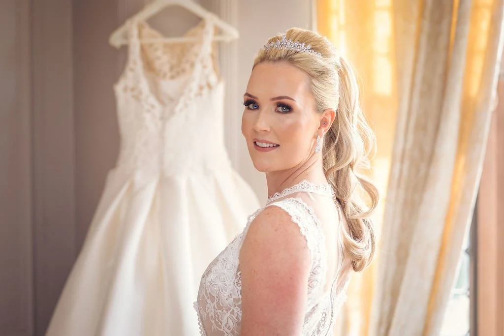 A smiling blonde bride in lace gown with a tiara, standing next to her wedding dress hanging on a wall.