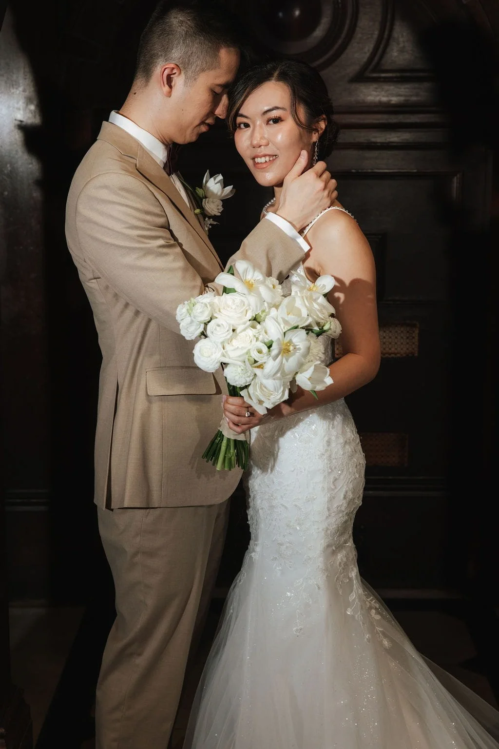 A bride and groom pose closely at their wedding, with the groom gently touching the bride's face. The bride is holding a bouquet of white flowers and is dressed in a lace wedding gown, while the groom is in a beige suit with a boutonniere.