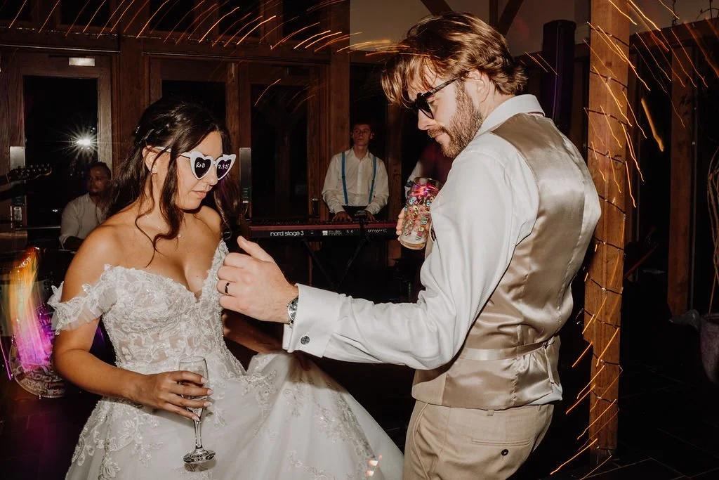 A woman in a wedding dress and sunglasses with heart shapes, and a man in a tuxedo vest, are dancing at a wedding reception. The woman holds a glass of champagne, and there is a live band playing in the background.