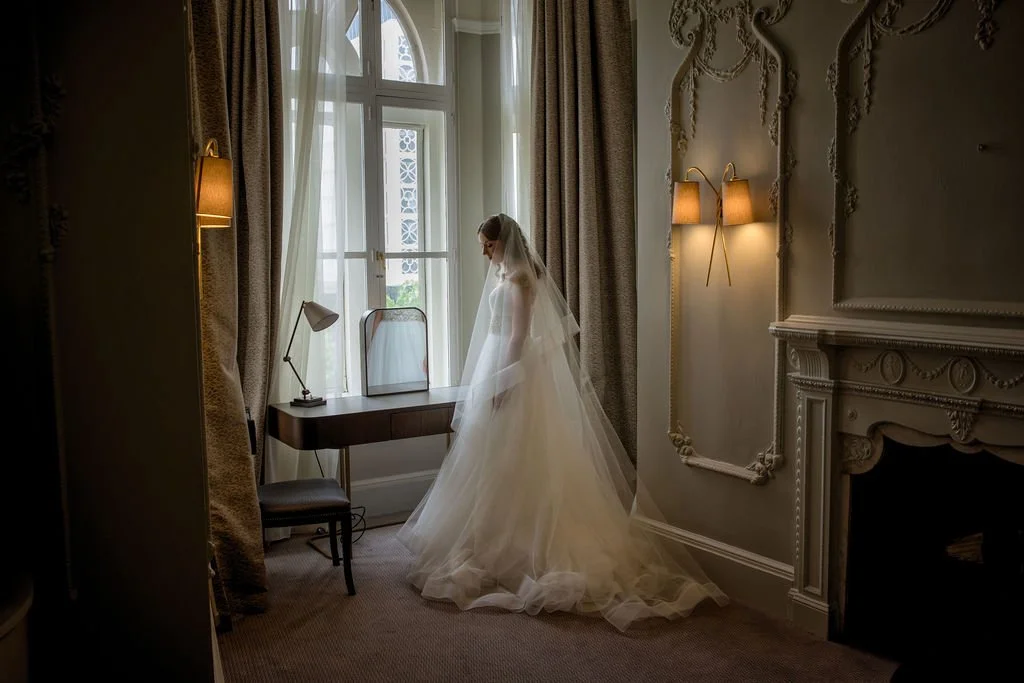 Bride in wedding dress standing near window in a decorated room with ornate wall details and fireplace.