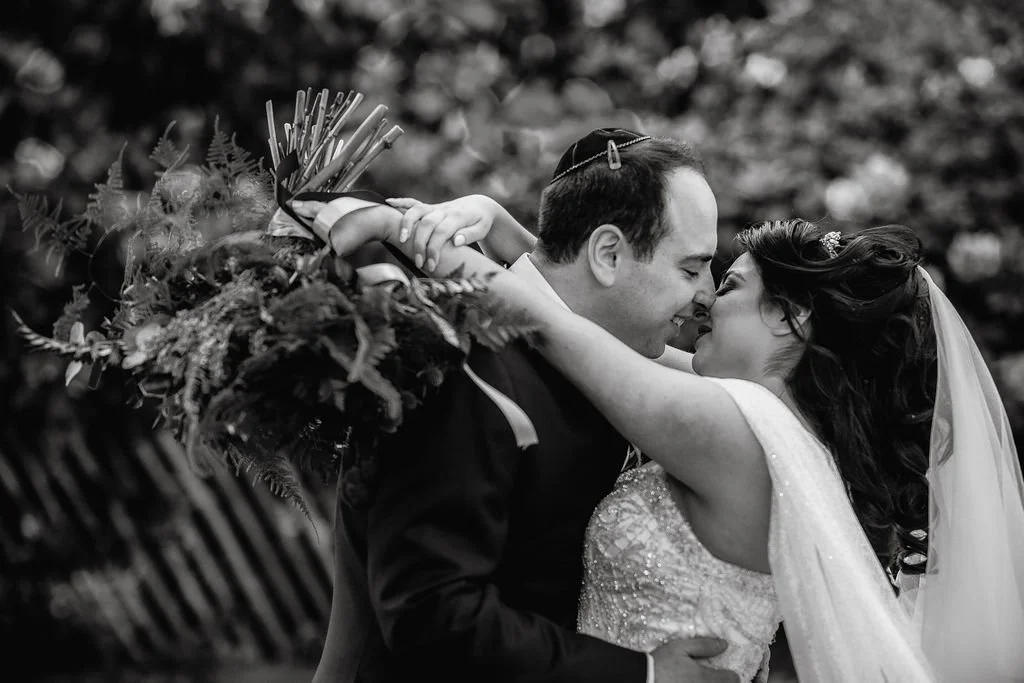 A black and white photograph of a bride and groom embracing and touching foreheads, with the bride holding a bouquet of flowers.