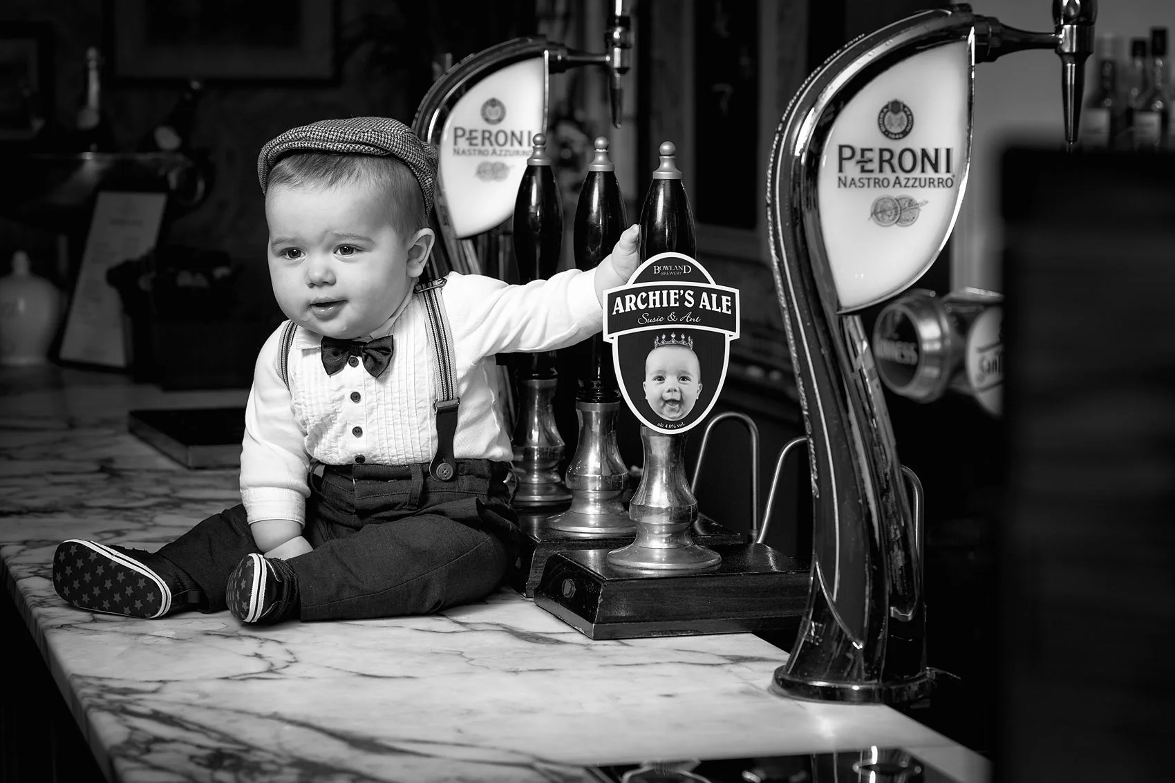 A young boy dressed in a tuxedo with a bowtie and suspenders sitting on a marble countertop, holding a sign that reads 'Archie's Ale' with a picture of his face, in a bar setting with beer taps in the background.