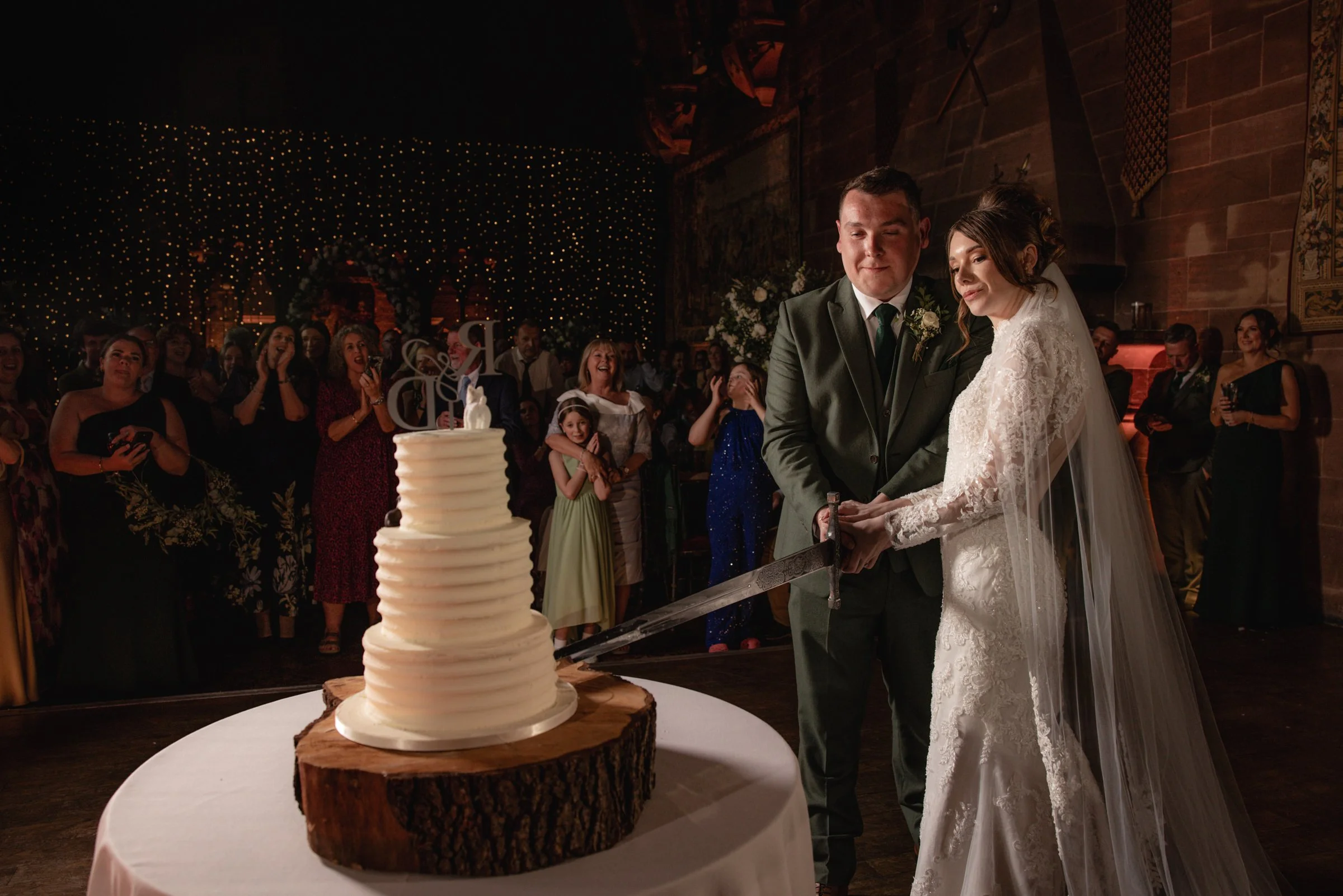 Wedding ceremony with bride and groom cutting a wedding cake surrounded by guests in a dimly lit venue with string lights and decorative banners.