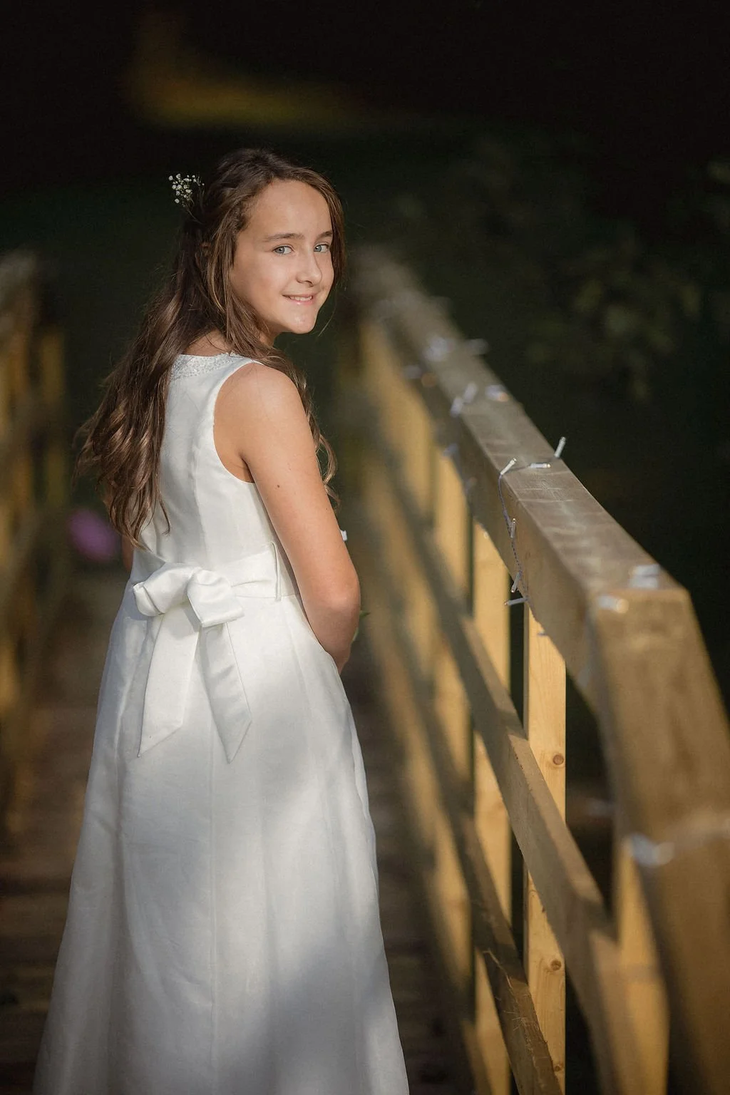 A young girl in a white sleeveless dress with a bow in her long, wavy brown hair, standing on a wooden dock at night, looking over her shoulder at the camera with a slight smile.