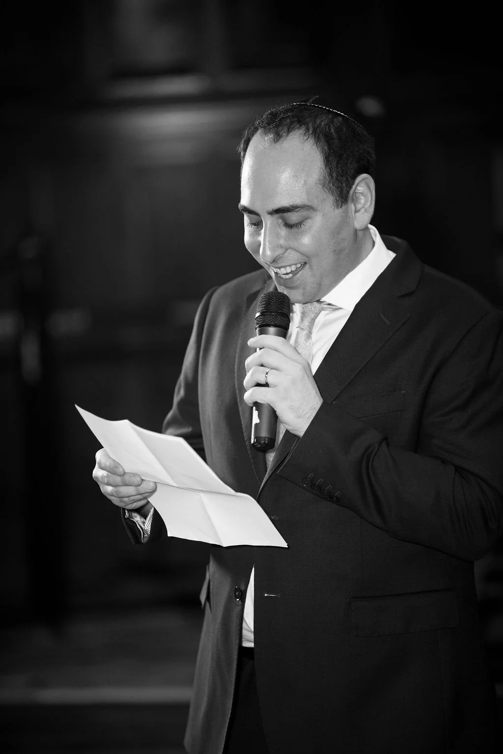 A man in a suit speaking into a microphone and reading from a piece of paper at an indoor event.