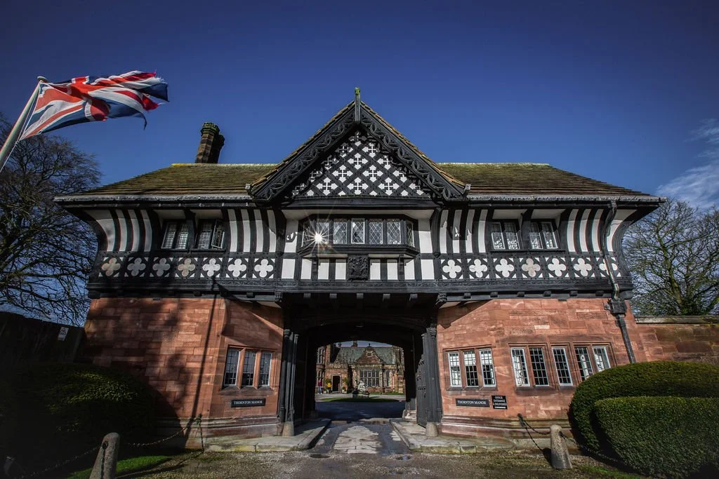 Historic black and white timber-framed building with a stone base and an archway entrance, located in a park-like setting with bushes and trees, featuring a British flag flying on the left.