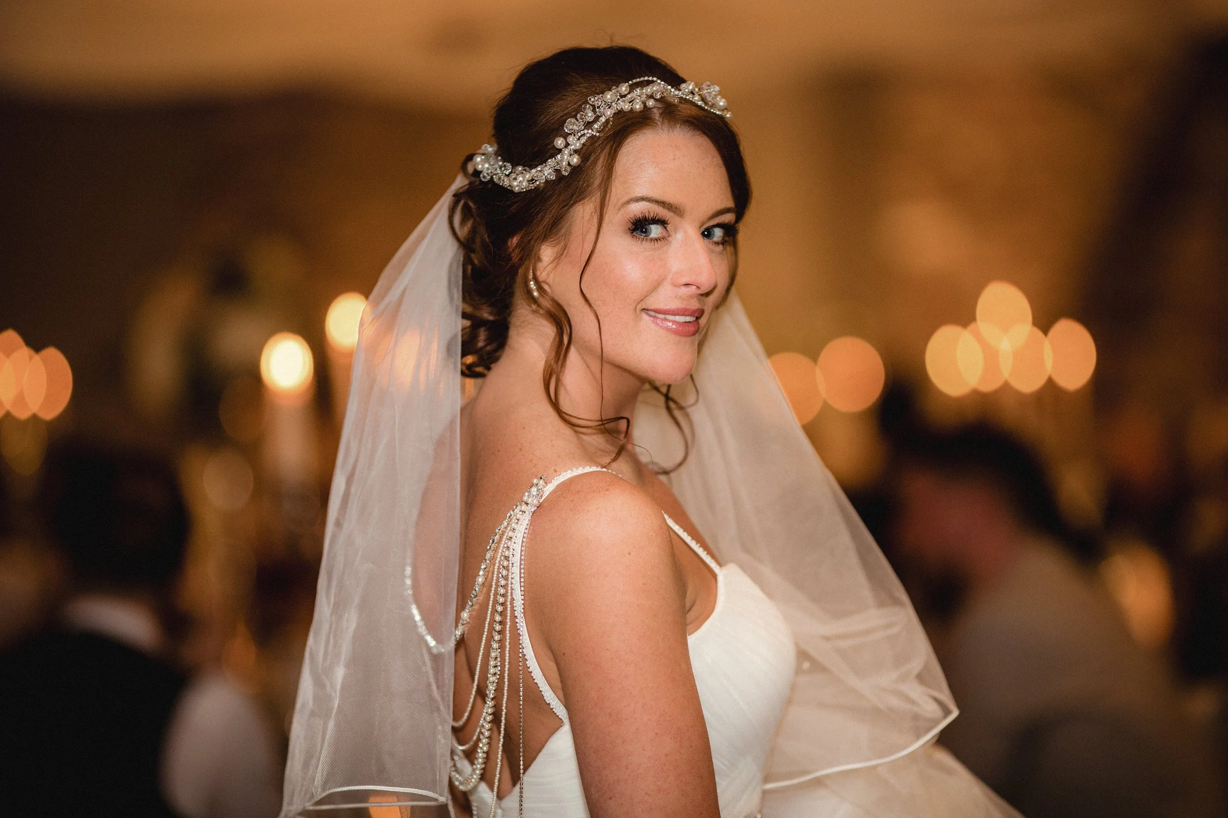 A bride with red hair styled in loose curls, wearing a white wedding gown with pearl and rhinestone straps, a sparkling rhinestone tiara, and a sheer veil, smiling softly at the camera in a warmly lit venue.