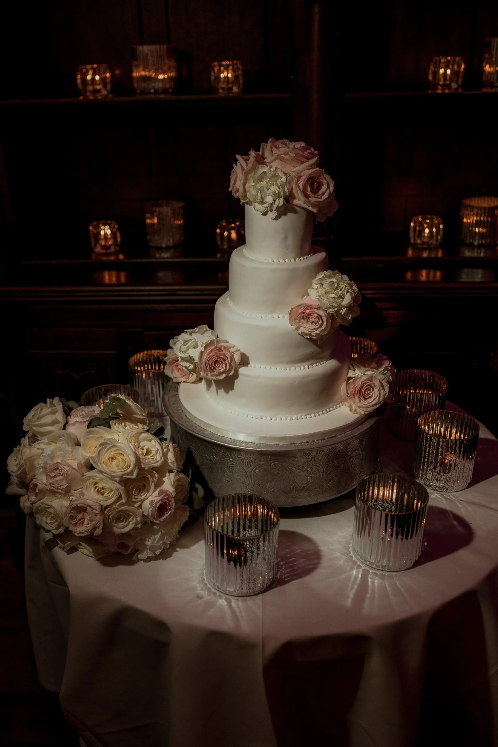 A four-tier white wedding cake decorated with pink and white roses on a table with a floral bouquet and silver candlesticks, dimly lit for a celebration.