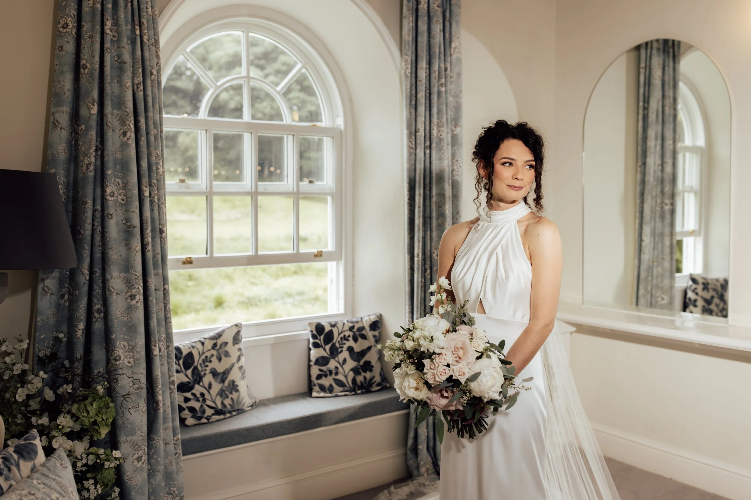 A woman in a wedding dress holding a bouquet of flowers inside a room with large arched windows, floral curtains, and a cushioned window seat with patterned pillows.