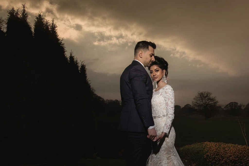 A bride and groom standing close together outdoors at dusk, with the sky and trees in the background.