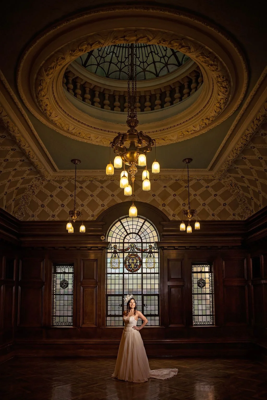 A woman in a white wedding dress standing in a grand, historic room with dark wood paneling, stained glass windows, and elegant chandelier lighting.