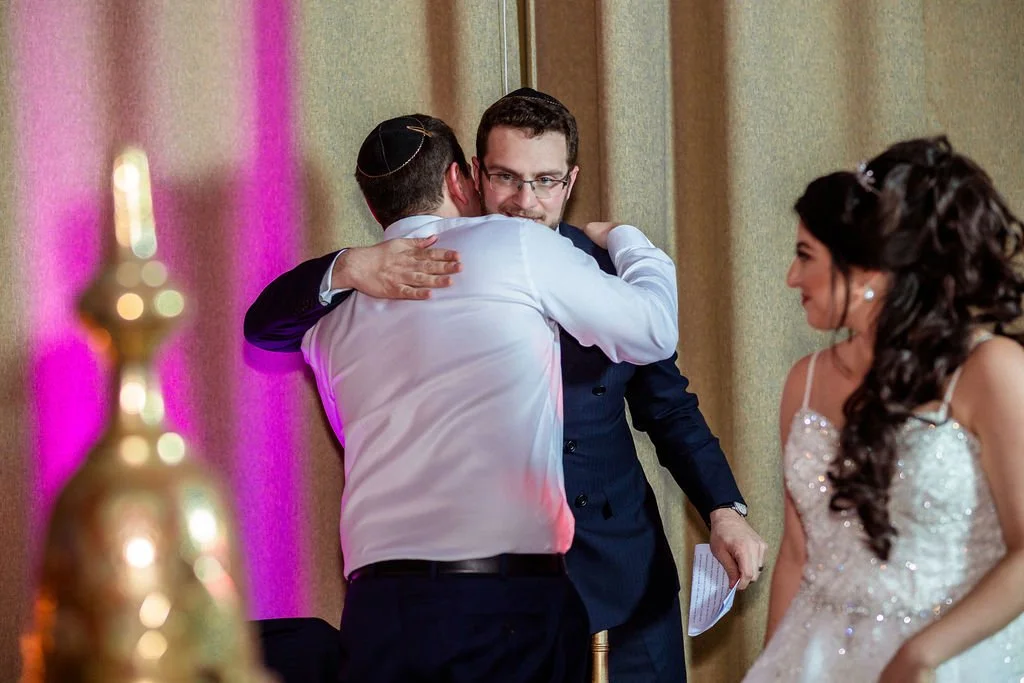 Two men hugging, one in a white shirt and the other in a dark suit, at a wedding reception; woman in a white wedding dress watching and smiling.