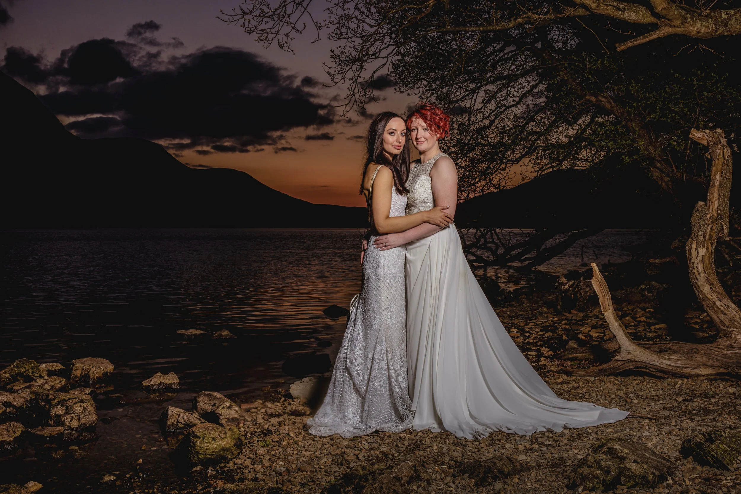 Two brides in white wedding dresses embrace during sunset at a lake distict lake with mountains in the backgrounf