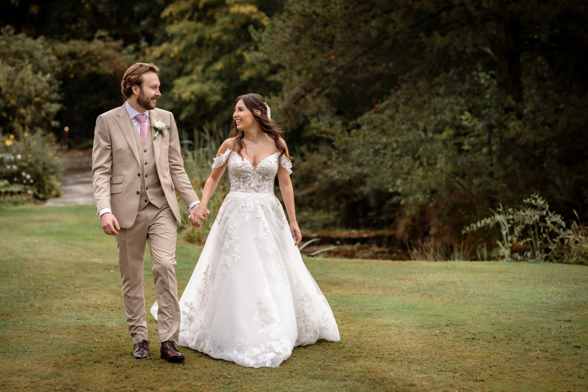 A bride and groom walking hand in hand on a grassy area outdoors, surrounded by trees, during their wedding celebration.