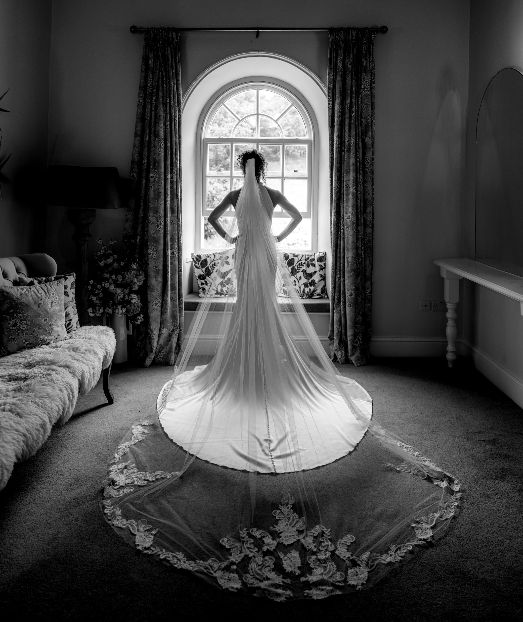 A woman in a wedding dress standing with her back to the camera, facing a large arched window with curtains, in a room with furniture and a carpeted floor.