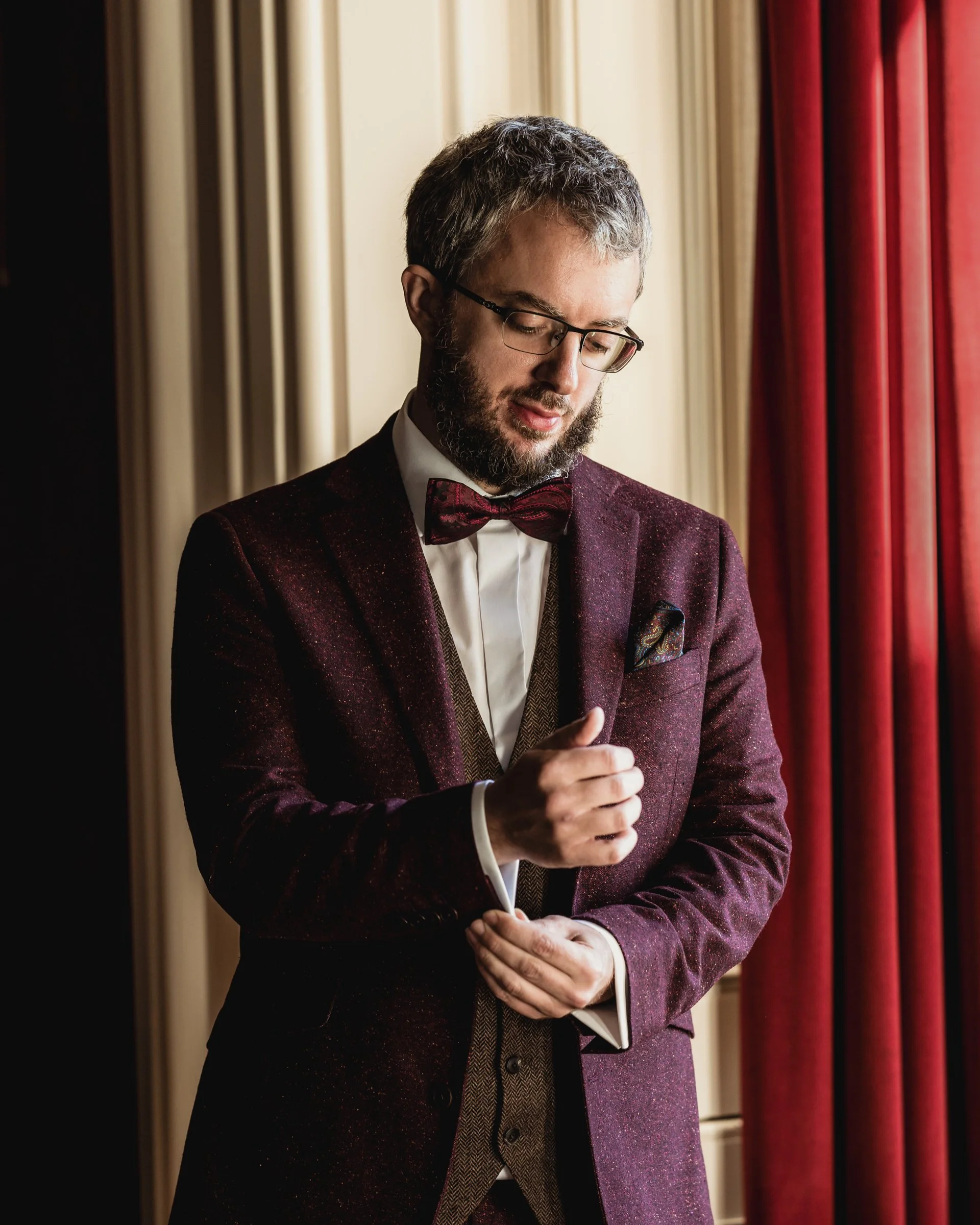A man with glasses, a beard, and gray hair is dressed in a maroon suit with a bowtie. He is adjusting his sleeve near a window with red curtains, and cream-colored panels are in the background.