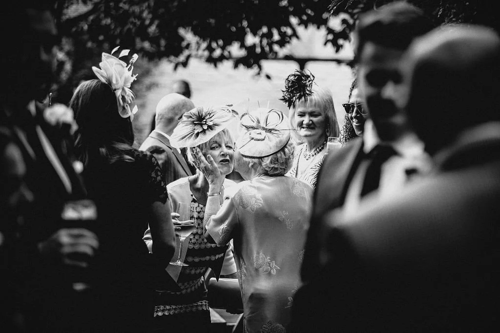 Group of people in formal attire, engaging in conversation outdoors during daytime under trees, some wearing decorative hats.
