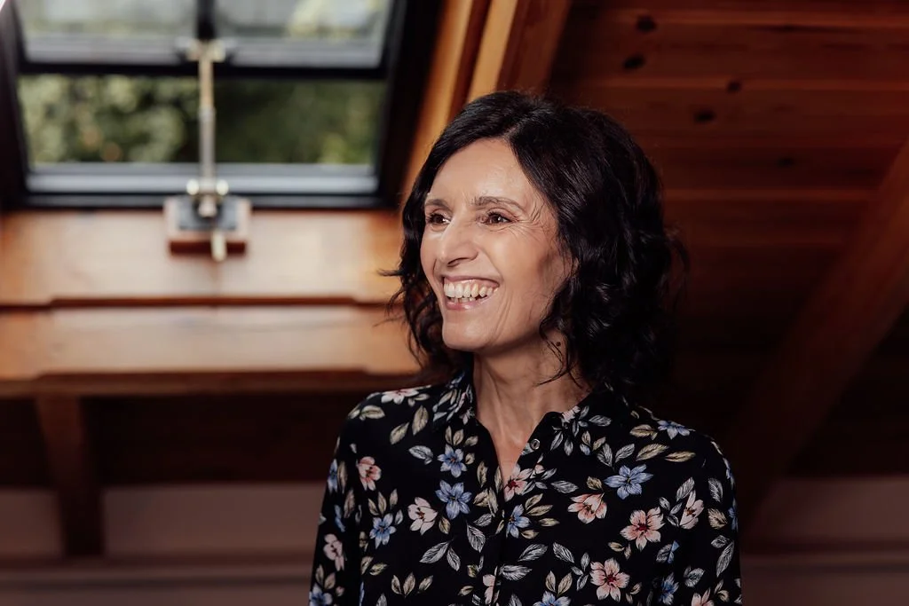 A woman with black, curly hair smiling, wearing a black blouse with a floral pattern, standing in a room with a wooden ceiling and a skylight window.