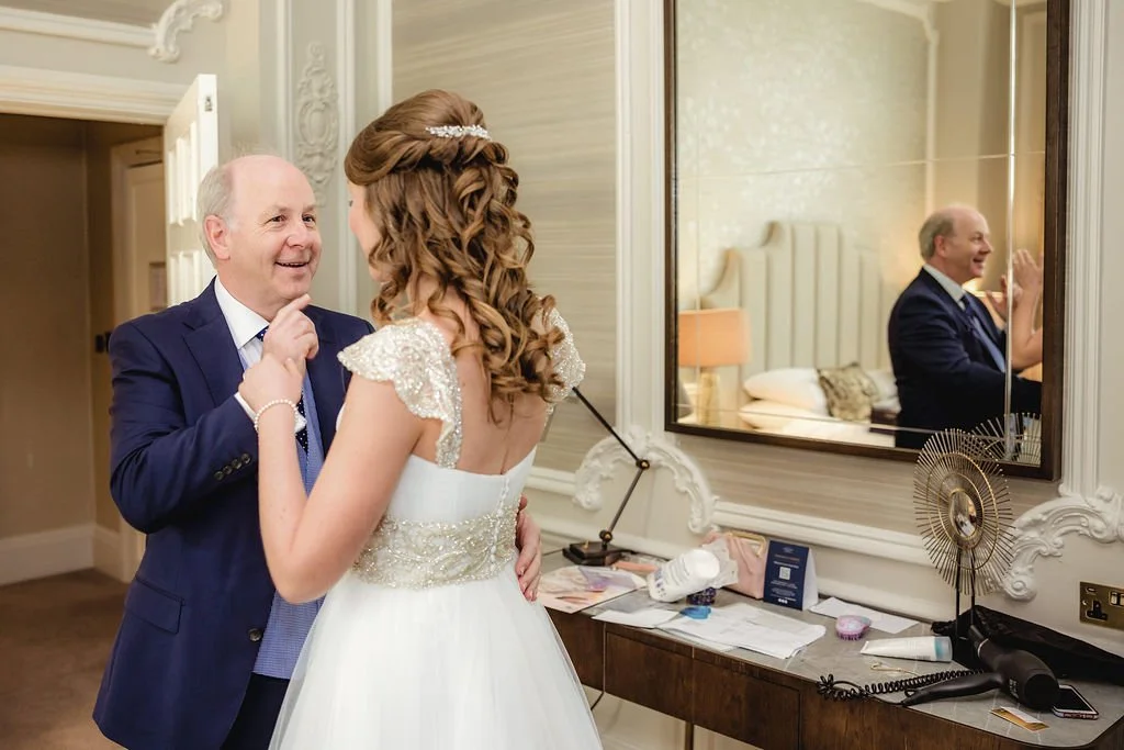 A bride in a white wedding dress with beaded shoulders talking to an older man in a blue suit inside a hotel room, with a mirror reflecting their faces.