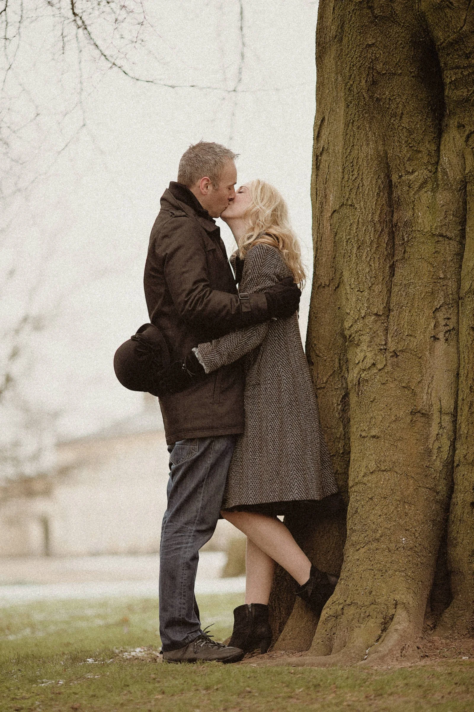 A couple sharing a kiss outdoors near a large tree, dressed in winter clothing.