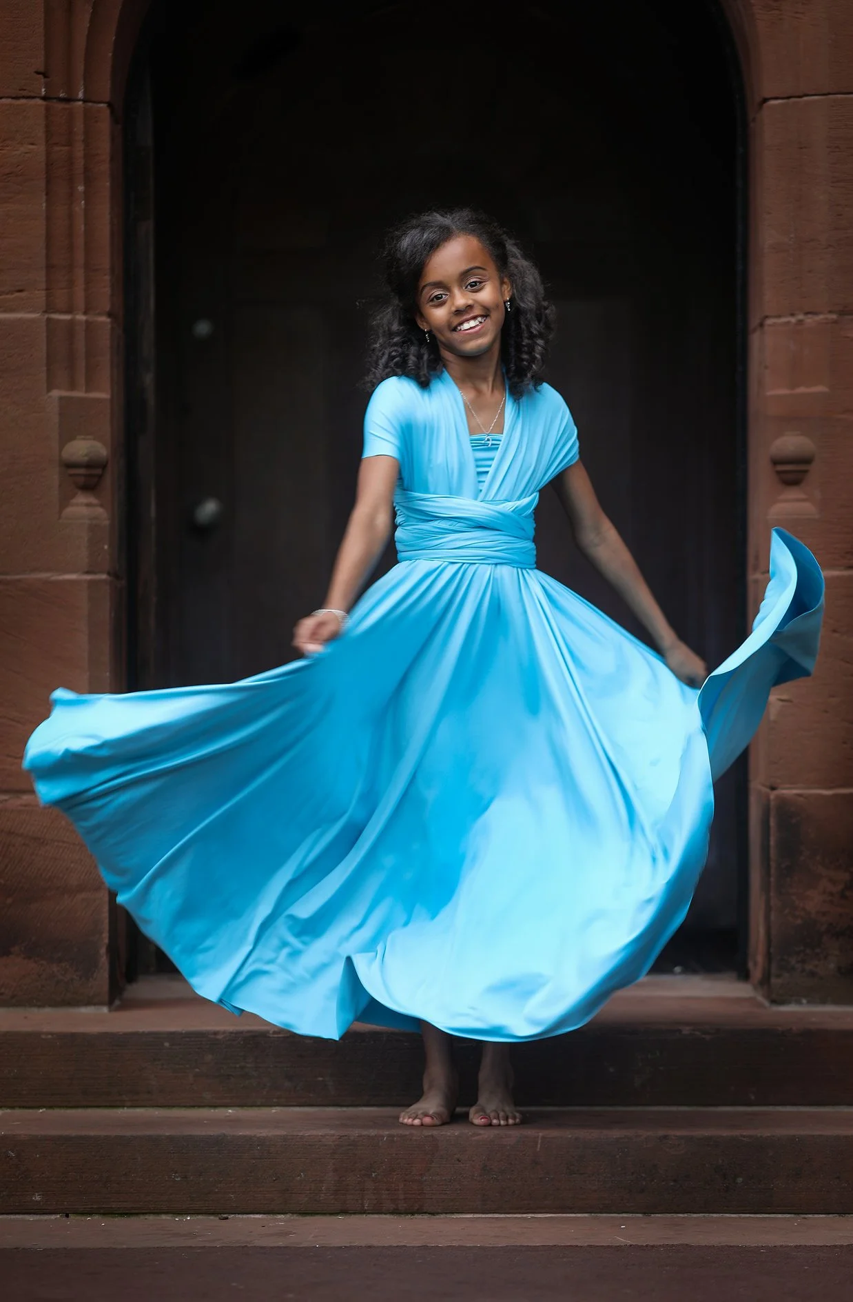A young girl in a bright blue dress standing on wooden steps, smiling and twirling, with a dark background and red brick arch framing her.