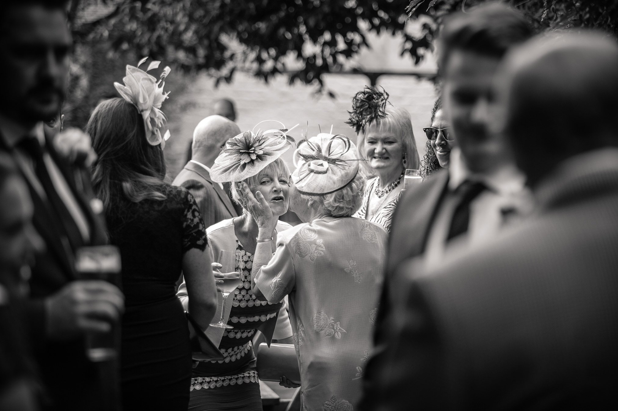 Guests gather and chat outside and two old ladies kiss and greet each other in the cetre of the scene.